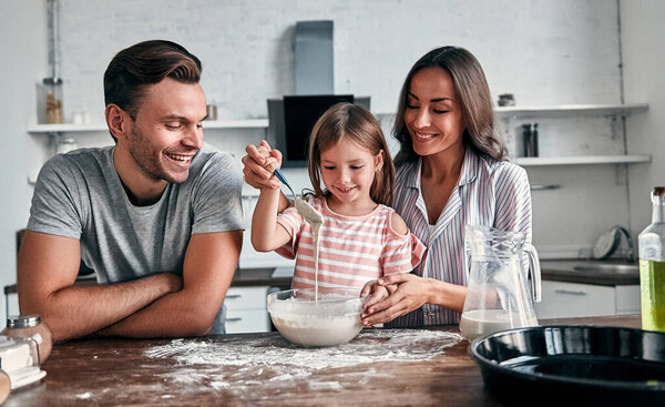 Cute little girl and her beautiful parents are knead flour for baking and smile while cooking in the kitchen.