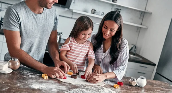 stock image Cute little girl and her beautiful parents are cookie cut out from dough and smile. Happy family in the kitchen preparing baked goods.