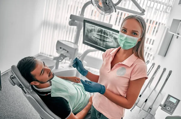 Female dentist in mask examining a patient with tools in dental clinic. Doctor doing dental treatment on man's teeth in the dentists chair.