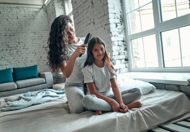 Güzel genç anne, küçük tatlı kızının saçlarını tarıyor. Hem pijamalarıyla, hem de yatakta oturmuş gülümsüyor.