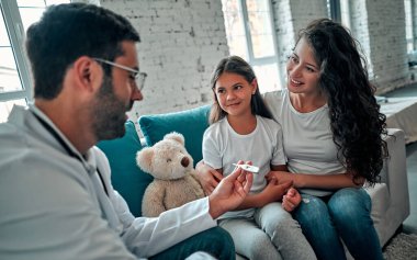 Güzel genç anne ve küçük kızı evde. Doktor küçük hastayı termometre kullanarak muayene ediyor..