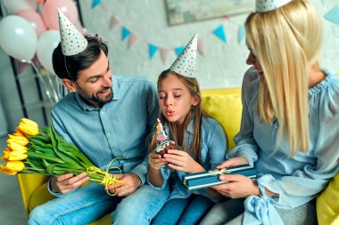 İyi ki doğdun! Mutlu ebeveynler ve kızları pastaya mum üflüyorlar. Küçük kızın doğum günü için aile partisi, oda balonlarla süslenmiş. Baba ve anne kızını tebrik eder ve ona bir hediye ve çiçek lalesi verir..