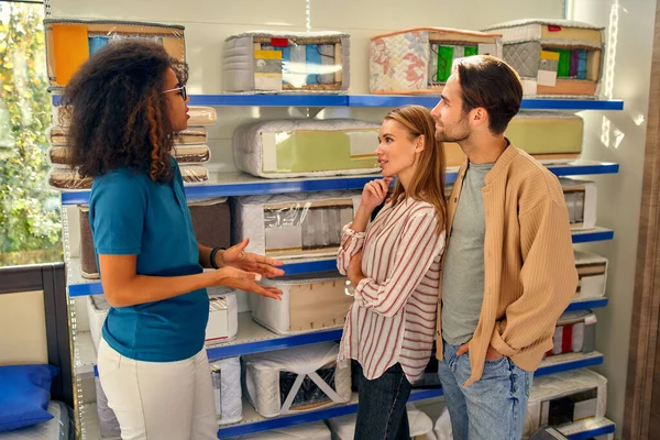 An African American woman salesperson consultant consults a young couple by showing samples of mattresses on shelves in a bed, mattress and pillow store.