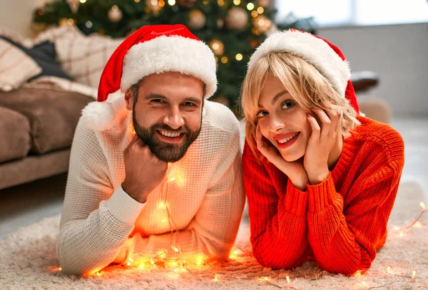 Mutlu Noeller ve mutlu yıllar! Noel Baba şapkalı genç bir çift Noel ağacının yanında yere uzanmış, tatili bekliyor..