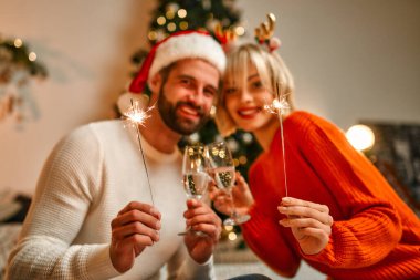 Mutlu Noeller ve mutlu yıllar! Noel Baba şapkalı ve ren geyiği boynuzları takmış bir çift Noel ağacının yanında, ellerinde maytaplar ve bir bardak şampanyayla tatil için bekliyorlar..