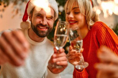 Mutlu Noeller ve mutlu yıllar! Noel Baba şapkalı ve ren geyiği boynuzları takmış bir çift Noel ağacının yanında, ellerinde maytaplar ve bir bardak şampanyayla tatil için bekliyorlar..