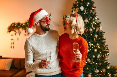 Mutlu Noeller ve mutlu yıllar! Noel Baba şapkalı ve boynuzlu komik geyik boynuzları takmış bir çift Noel ağacının yanında oturmuş, ellerinde şampanya kadehleriyle bayramı bekliyorlar..