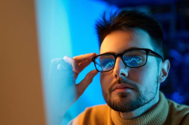 Beyaz tenli, gözlüklü genç bir satıcının ofiste çalışırken monitörlere grafiklerle bakması. Borsa, insanlar, iş konsepti.