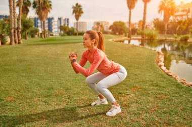 Spor giyimli genç, narin bir kadın çimlerin üzerinde palmiye ağaçlarının arka planında çömelmiş egzersiz yapıyor. Spor ve Eğlence..