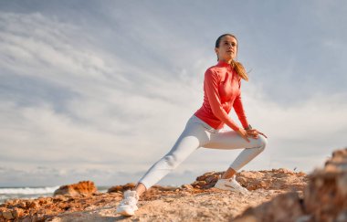 Spor giyimli, narin genç bir kadın denizin arka planında taşların üzerinde dikilirken egzersiz yapıyor. Spor ve Eğlence..