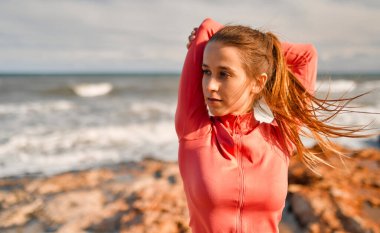 Spor giyimli, narin genç bir kadın denizin arka planında taşların üzerinde dikilirken egzersiz yapıyor. Spor ve Eğlence..