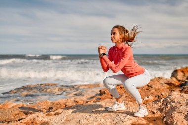 Spor giyimli, narin genç bir kadın denizin arka planında taşların üzerinde dikilirken egzersiz yapıyor. Spor ve Eğlence..