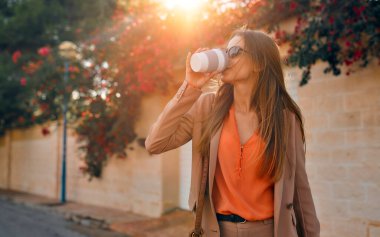 Takım elbiseli, güneş gözlüklü genç bir iş kadını ya da öğrenci çiçeklerle dolu bir kasabanın sokaklarında yürürken gülümser ve kahve içer..