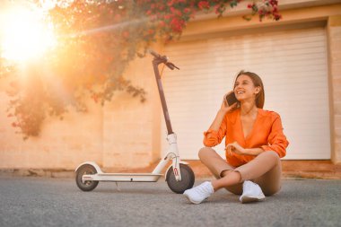 Elektrikli scooterlı genç bayan iş kadını ya da öğrenci çiçeklerle dolu bir kasabanın caddesinde otururken telefonla konuşuyor..