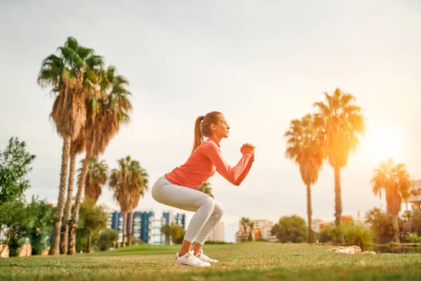 Spor giyimli genç, narin bir kadın çimlerin üzerinde palmiye ağaçlarının arka planında çömelmiş egzersiz yapıyor. Spor ve Eğlence..