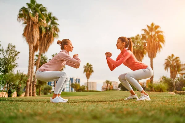 Spor giyimli genç, narin kadınlar bahçede palmiye ağaçlarının arka planında çömelme egzersizi yapıyorlar. Spor ve Eğlence..