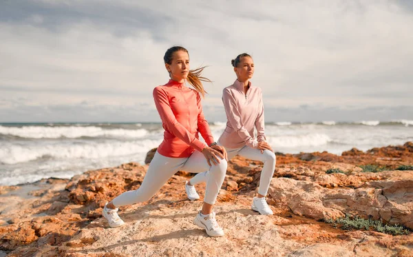 Spor giyimli narin genç kadınlar denizin arka planında taşların üzerinde dururken egzersiz yapıyorlar. Spor ve Eğlence..