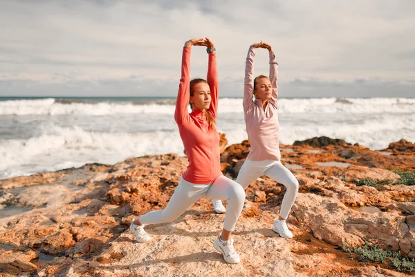 Spor giyimli narin genç kadınlar denizin arka planında taşların üzerinde dururken egzersiz yapıyorlar. Spor ve Eğlence..