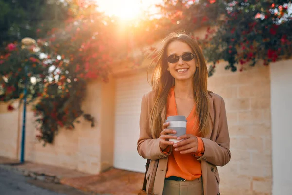 Takım elbiseli, güneş gözlüklü genç bir iş kadını ya da öğrenci çiçeklerle dolu bir kasabanın sokaklarında yürürken gülümser ve kahve içer..