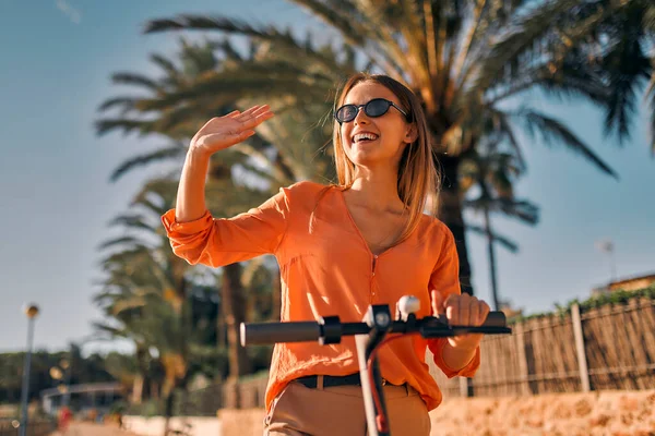 Genç stil sahibi kadın iş kadını ya da öğrenci, sahilde palmiye ağaçlarının ve dalgaların arkasına elektrikli scooter sürüyor..