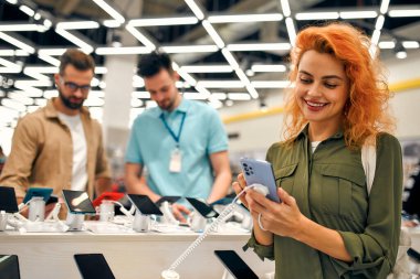 Kızıl saçlı tatlı kadın ev aletleri, elektronik aletler ve aletler mağazasından yeni akıllı telefon seçiyor. Arka planda, satıcı alıcıya tavsiye eder. İndirim Günü.