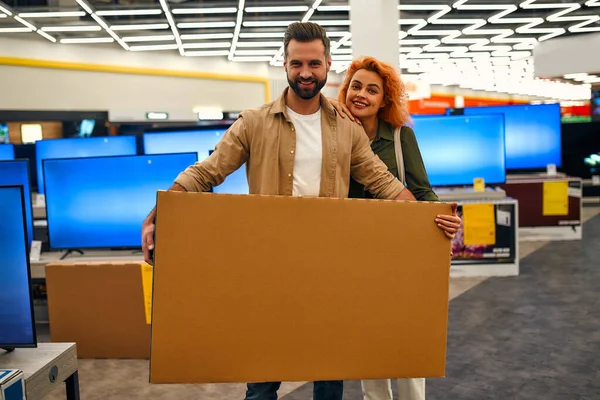 Happy young couple bought new TV, holding box with purchase in store of household appliances, electronics and gadgets. Buying new TV, sale day.