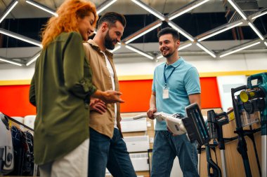 Genç çift ev aletleri ve elektronik mağazasından yeni elektrikli süpürge alıyor. Satıcı, alıcılara ekipman alımında seçim yapmalarını tavsiye eder..