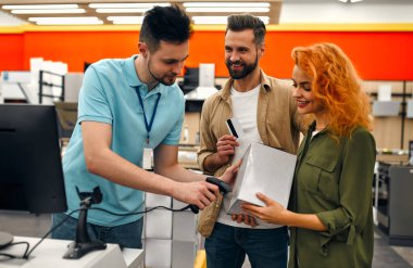 Bir kadın ve bir adam kasada, ev aletleri ve elektronik mağazasında bir kutu ve kredi kartı tutuyor. Erkek satıcı ürün kutusundaki kodu tarayıcıyla okuyor..