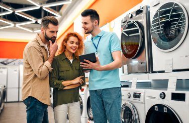 Güzel kızıl saçlı bir kız ve sakallı bir adam ev aletleri ve elektronik mağazasından çamaşır makinesi alıyorlar. Erkek bir satıcı, çiftlere evleri için yeni aletler seçmelerine yardım eder..