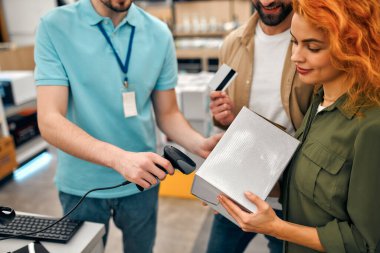 Bir kadın ve bir adam kasada, ev aletleri ve elektronik mağazasında bir kutu ve kredi kartı tutuyor. Erkek satıcı ürün kutusundaki kodu tarayıcıyla okuyor..