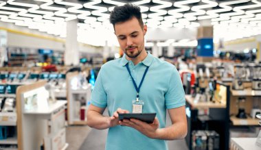 Üniformalı başarılı bir satış asistanı elinde tabletle evin arka planındaki elektronik eşya mağazasında çalışıyor..