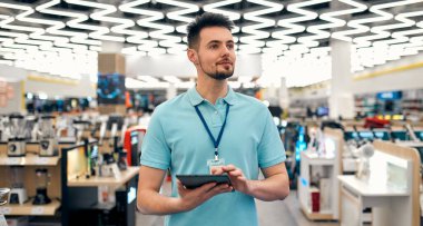 Üniformalı başarılı bir satış asistanı elinde tabletle evin arka planındaki elektronik eşya mağazasında çalışıyor..