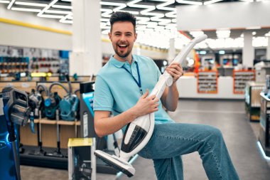 Üniformalı başarılı bir satış asistanı elektrikli süpürge gibi arka planda bir ev aletleri ve elektronik mağazasında gitar çalarken eğleniyor..