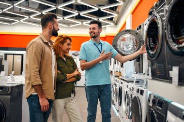 Güzel kızıl saçlı bir kız ve sakallı bir adam ev aletleri ve elektronik mağazasından çamaşır makinesi alıyorlar. Erkek bir satıcı, çiftlere evleri için yeni aletler seçmelerine yardım eder..
