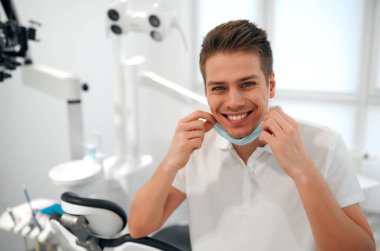 Beyaz üniformalı, kar beyazı bir gülümseme ve diş hekimliği bürosundaki bir sandalyede oturan koruyucu tıbbi maskeli genç bir dişçinin portresi..