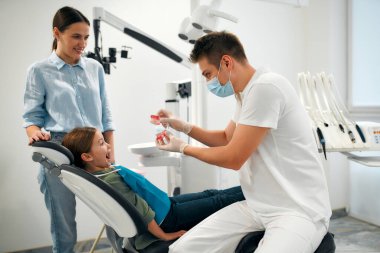 Annesi dişçi koltuğunda oturan tatlı bir kız çocuğu. Pediyatrik dişçi çene modelini gösterir ve hastaya diş bakımını açıklar.