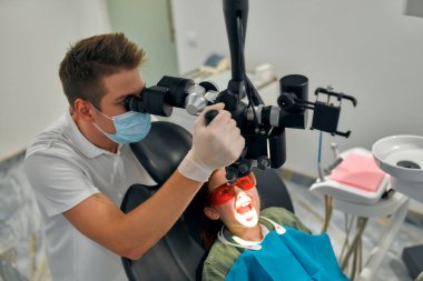 Erkek bir dişçi dişlerine bakan güzel bir kızı dişçi muayenehanesinde profesyonel bir mikroskopla muayene ediyor. Diş işleten mikroskop. Diş optiği.