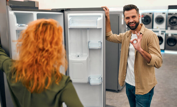 Молодая пара выбирает новый холодильник в магазине бытовой техники и электроники. Мужчина с бородой показывает нормальный жест, держа дверь холодильника..