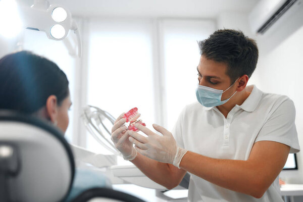 Young dentist in white uniform and protective medical mask showing jaw model with braces to female patient in dentistry office with modern equipment.