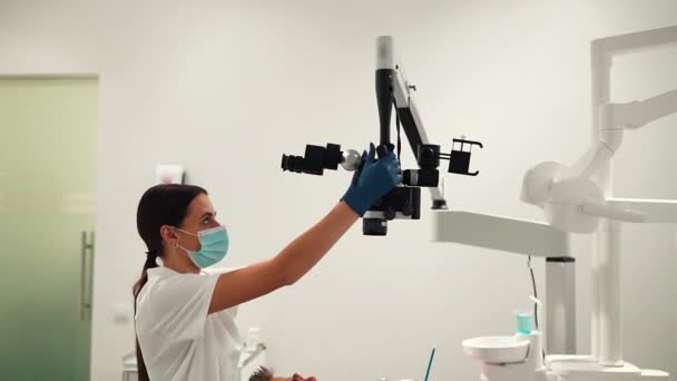 女性歯科医は 歯科手術室でプロの顕微鏡で歯を見ている若い患者を調べます 歯科手術用顕微鏡 歯科光学 — ストック動画