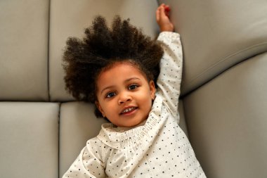 Şirin Afro-Amerikalı kıvırcık saçlı kız bebek evdeki oturma odasındaki kanepede uzanıyor..