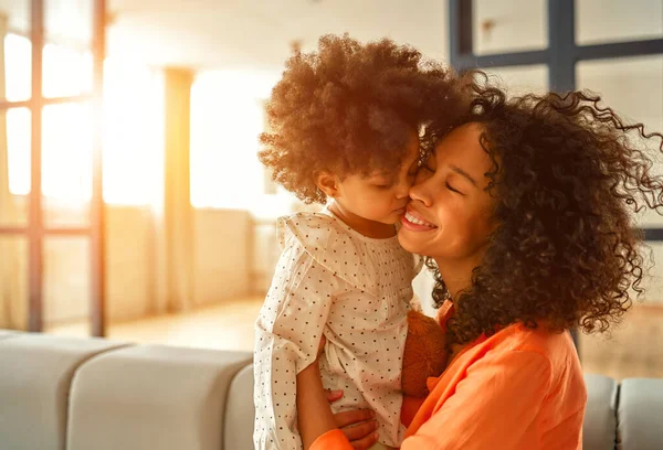 アフリカ系アメリカ人女性とともに彼女の小さな娘とともに巻き毛座っていますソファと優しく抱擁で自宅のリビングルーム — ストック写真
