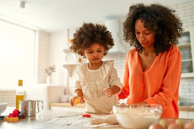 Küçük kızı kıvırcık saçlı Afro-Amerikalı kadın mutfakta hamur işi pişiriyor ve eğleniyor. Anne ve kızı birlikte yemek yapıyor. Kurabiye kesiciyle kurabiye kesen çocuk.
