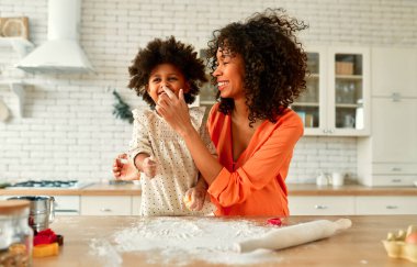 Küçük kızı kıvırcık saçlı Afro-Amerikalı kadın mutfakta hamur işi pişiriyor ve eğleniyor. Anne ve kızı birlikte yemek pişirip birbirlerini un ile kirletiyorlar..