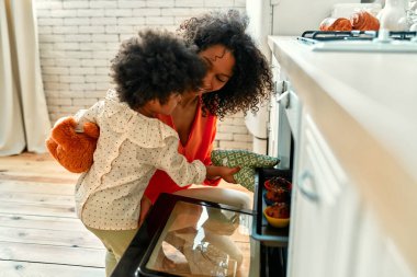 Kıvırcık saçlı küçük bir kızı olan Afro-Amerikalı bir kadın fırından fırında pişmiş keklerle birlikte bir yemek tepsisi çıkarıyor. Anne ve kızı mutfakta kek pişiriyor..