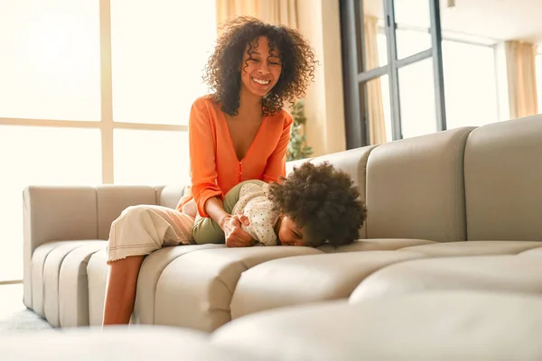 アフリカ系アメリカ人女性とともに彼女の小さな娘とともに巻き毛で座っていますザソファと楽しいですリビングルームに自宅 — ストック写真