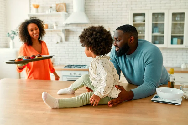 ふわふわの巻き毛の小さな娘を持つアフリカ系アメリカ人の家族は 台所で一緒にカップケーキを作りました お母さんは 既製のマフィンとベーキングシートを運ぶ — ストック写真