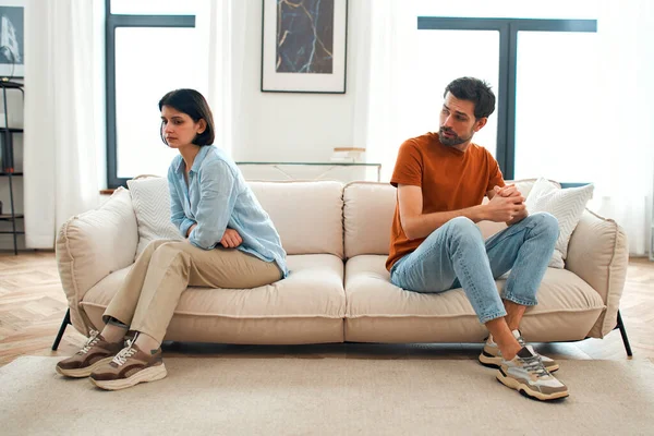 Unhappy sad couple sitting apart on couch in living room after quarrel, frustrated man and woman turning their back ignoring having conflict at home, can not find compromise, misunderstanding concept.