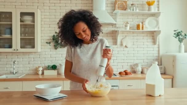 かわいいですアフリカ系アメリカ人女性とともにカールふわふわの髪は 彼女のキッチンで自宅で甘いベーキングクッキーのための生地を準備しながら ボウルにミキサーで卵を泡立てています — ストック動画