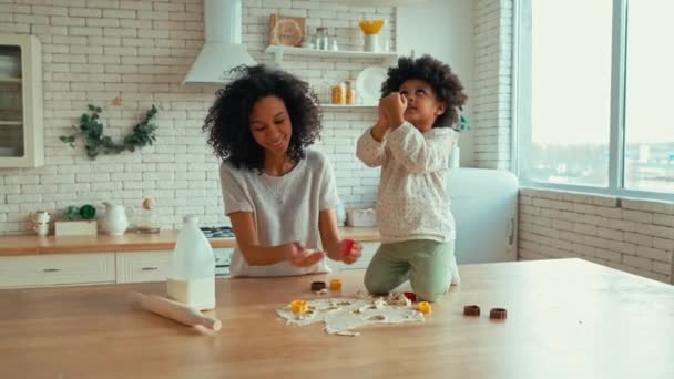 キッチンで楽しさと料理のペストリーを持っている巻きふわふわの髪を持つ彼女の小さな娘とアフリカ系アメリカ人の女性 母と娘が一緒に料理をする クッキーカッターでクッキーを切り取る子 — ストック動画
